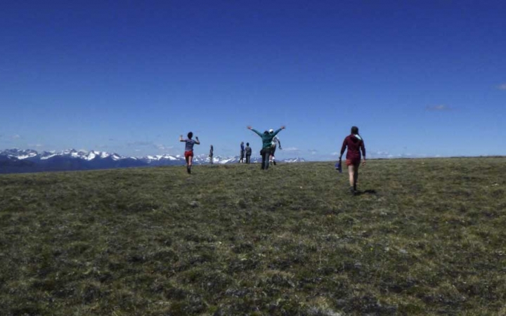 A group of people hike across a grassy meadow toward snow-capped mountains in the background